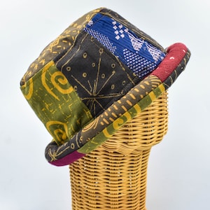 Może przedstawiać: Patchworkowy kapelusz typu bucket z różnorodnych wzorzystych tkanin w odcieniach zieleni, niebieskiego, czerni i złota. Rondo wykończone bordową tkaniną. Kapelusz jest prezentowany na wiklinowej głowie.