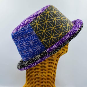 Può includere: Un cappello a secchiello con un design patchwork di motivi geometrici in viola, blu, giallo e nero. La corona e la tesa mostrano sezioni distinte con motivi floreali e stellati ripetuti, creando un accessorio tessile vivace e colorato.