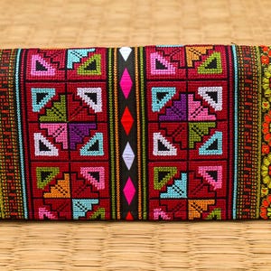 Könnte beinhalten: Rechteckige, bestickte Clutch mit einem lebendigen, geometrischen Muster. Das zentrale Design zeigt Quadrate mit bunten Dreiecken und Rauten, umrahmt von floralen und gestreiften Bordüren. Ein grüner Quastenanhänger und Holzperlen schmücken den Reißverschluss.