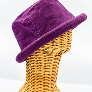 Puede incluir: Un sombrero de pana morado con una corona estructurada y un ala ligeramente curvada. El sombrero se exhibe sobre una cabeza de maniquí de mimbre sobre un fondo blanco. El color del sombrero es un tono morado intenso.