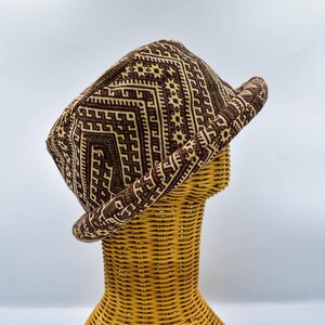 Op de afbeelding: Een bruin en beige patroonhoed met een geometrisch ontwerp. De hoed wordt getoond op een rieten hoofdvorm tegen een witte achtergrond. De rand van de hoed is opgerold en het patroon is zichtbaar op de boven- en zijkanten.