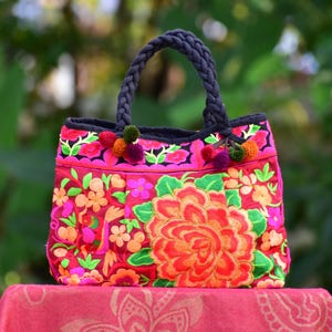 May include: A pink and red floral embroidered tote bag with black handles and colorful pom-poms.