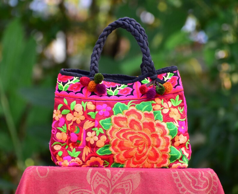 May include: A pink and red floral embroidered tote bag with black handles and colorful pom-poms.