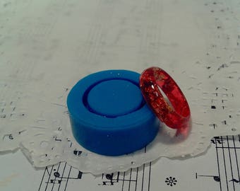 May include: A red resin ring with gold glitter embedded in the resin. The ring is sitting on a blue silicone ring mould.