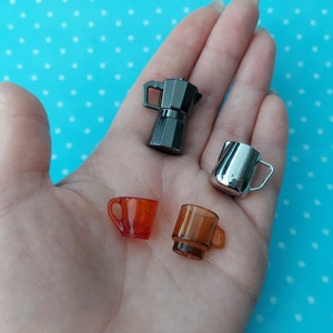 Pode incluir: Um conjunto de itens de cozinha em miniatura, incluindo uma cafeteira prateada, um jarro de creme prateado, uma xícara de vidro transparente e uma xícara de vidro laranja.