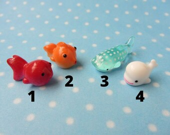 May include: Four small plastic figurines of sea creatures: a red fish, an orange fish, a blue whale shark, and a white whale. The figurines are numbered 1 through 4.