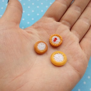 May include: Three miniature pies with different fillings. One pie has a red cherry filling, and the other two have white fillings. The pies are on a hand.