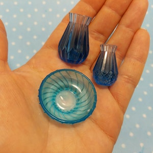 Puede incluir: Tres jarrones de vidrio azul en miniatura y un tazón de vidrio azul con un diseño de espiral. Los jarrones y el tazón están en una mano.