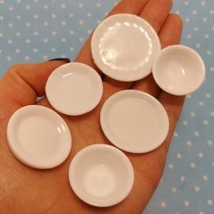 Op de afbeelding: Een set van zes witte plastic borden in verschillende maten, perfect voor een poppenhuis of miniatuurspel. De borden zijn in een hand gerangschikt, waardoor hun verschillende maten en vormen worden getoond.