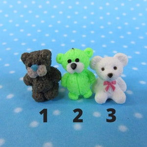 Puede incluir: Tres figuras de ositos de peluche en miniatura. Uno es gris oscuro con una nariz azul, el segundo es verde brillante y el tercero es blanco con un lazo rosa. Los osos están numerados 1, 2 y 3, sobre un fondo azul de lunares.