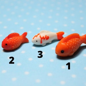 Puede incluir: Tres figuritas de peces en miniatura, dos son naranjas y una es blanca con marcas naranjas. Las figuritas están dispuestas sobre un fondo azul con lunares blancos.