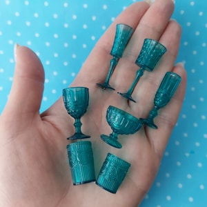 Puede incluir: Un juego de siete copas y vasos de vidrio azul verdoso con un patrón decorativo. Las copas tienen un tallo alto y delgado y una copa ancha. Los vasos son más bajos y tienen un lado recto.