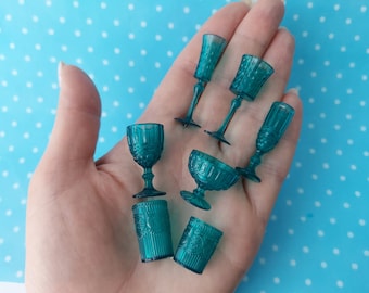 Puede incluir: Un juego de siete copas y vasos de vidrio azul verdoso con un patrón decorativo. Las copas tienen un tallo alto y delgado y una copa ancha. Los vasos son más bajos y tienen un lado recto.