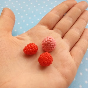 May include: Three small, realistic-looking raspberry-shaped polymer clay charms. Two are red and one is pink.