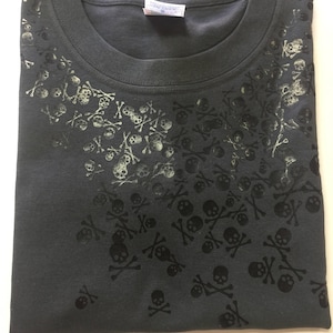 Könnte beinhalten: Ein dunkelgraues T-Shirt mit einem schwarz-weißen Totenkopf- und Knochenmuster. Das Muster ist auf der linken Seite des Shirts konzentriert und verblasst nach rechts.