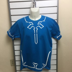 Puede incluir: Una camiseta azul con un diseño blanco de una espada y formas geométricas. El diseño está en la parte delantera de la camiseta y en las mangas.