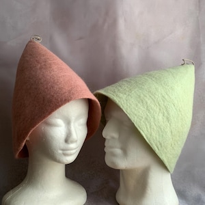 May include: Two felt hats, one in a soft pink and the other in a light green, are displayed on white mannequin heads. Each hat has a conical shape and a small, decorative silver loop at the top. The hats are made of a textured, felt material.