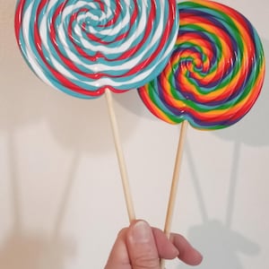 Peut inclure: Deux grandes sucettes avec des bâtonnets en bois. L'une présente un motif en spirale rouge, blanc et bleu. L'autre a un motif en spirale arc-en-ciel. Les sucettes sont tenues sur un fond blanc uni.