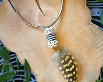 May include: A black and white patterned necklace with a gold metal chain and a feather pendant. The feather is brown and yellow with white spots.
