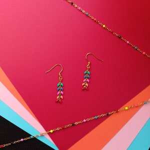 Pode incluir: Um par de brincos de argola dourados com contas coloridas penduradas na parte inferior. Os brincos estão sobre um fundo multicolorido.