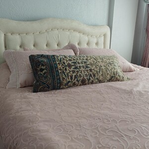 May include: A bed with a cream-colored, button-tufted headboard and a light pink bedspread. Decorative pillows in shades of pink and a long rectangular pillow with a floral pattern in green, blue, and beige complete the bedding ensemble.