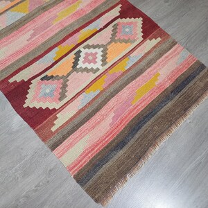 May include: A colorful handwoven rug with a geometric pattern in shades of red, pink, brown, and yellow. The rug has a fringed edge.