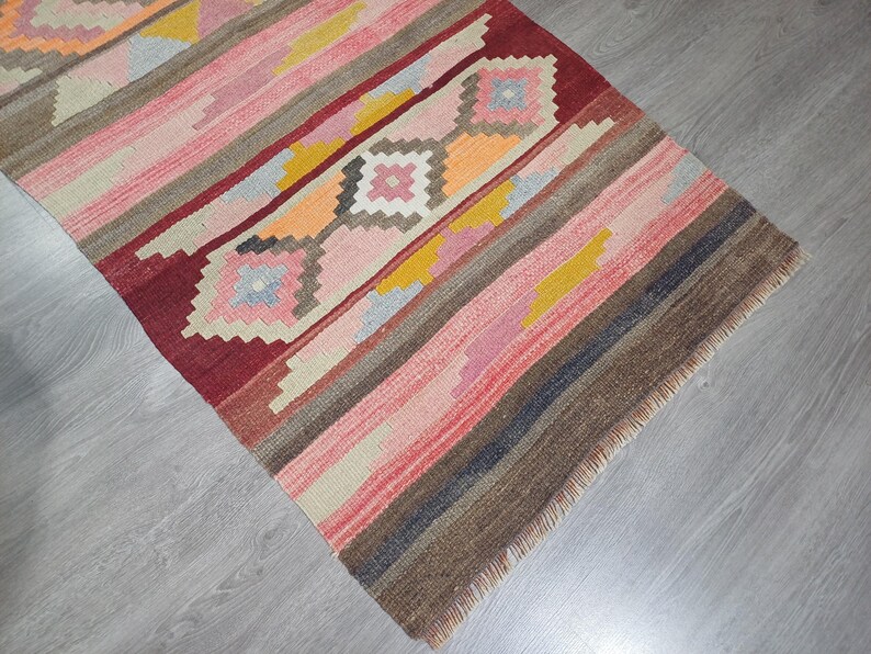 May include: A colorful handwoven rug with a geometric pattern in shades of red, pink, brown, and yellow. The rug has a fringed edge.