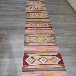 May include: A long, narrow rug with a colorful geometric pattern in shades of pink, red, yellow, blue, and brown. The rug has a traditional, handwoven look.