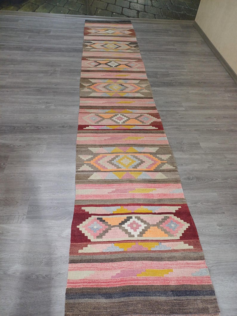 May include: A long, narrow rug with a colorful geometric pattern in shades of pink, red, yellow, blue, and brown. The rug has a traditional, handwoven look.