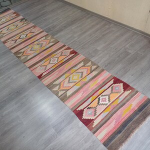 May include: A long, narrow rug with a geometric pattern in shades of pink, yellow, orange, and brown. The rug has a traditional, hand-woven look.