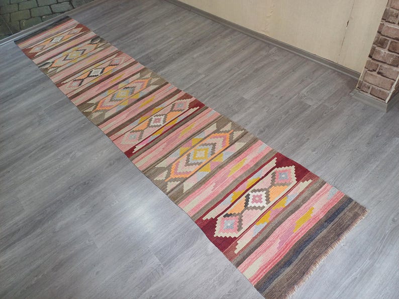 May include: A long, narrow rug with a geometric pattern in shades of pink, yellow, orange, and brown. The rug has a traditional, hand-woven look.