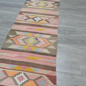May include: A long, narrow rug with a geometric pattern in shades of pink, brown, and yellow. The rug has a traditional, hand-woven look.