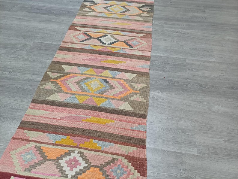 May include: A long, narrow rug with a geometric pattern in shades of pink, brown, and yellow. The rug has a traditional, hand-woven look.
