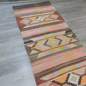 May include: A long, narrow rug with a colorful geometric pattern. The rug is woven with shades of pink, orange, brown, and gray. The pattern features repeating diamond shapes and stripes.