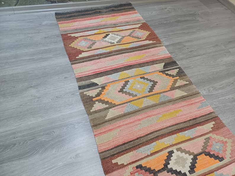 May include: A long, narrow rug with a colorful geometric pattern. The rug is woven with shades of pink, orange, brown, and gray. The pattern features repeating diamond shapes and stripes.
