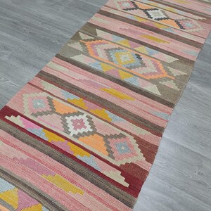 May include: A long, narrow rug with a pink, brown, and yellow geometric pattern. The rug has a traditional, hand-woven look.