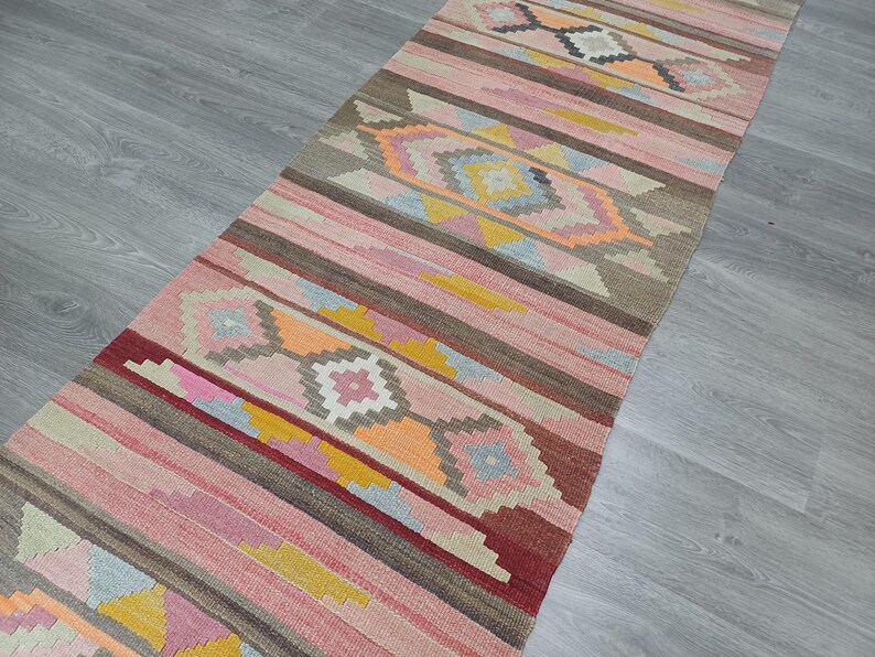 May include: A long, narrow rug with a pink, brown, and yellow geometric pattern. The rug has a traditional, hand-woven look.