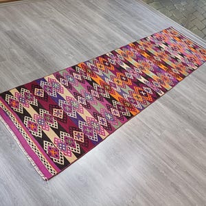 Puede incluir: Una alfombra larga y colorida con un patrón geométrico en tonos de rosa, morado, naranja y negro. La alfombra tiene un patrón de diamante con un diseño de cruz central.