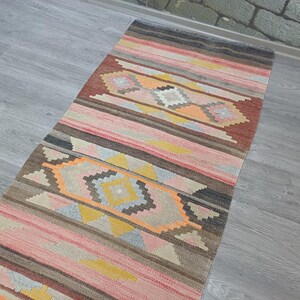 May include: A long, narrow rug with a colorful geometric pattern in shades of pink, orange, yellow, brown, and gray. The rug has a traditional, handwoven look.