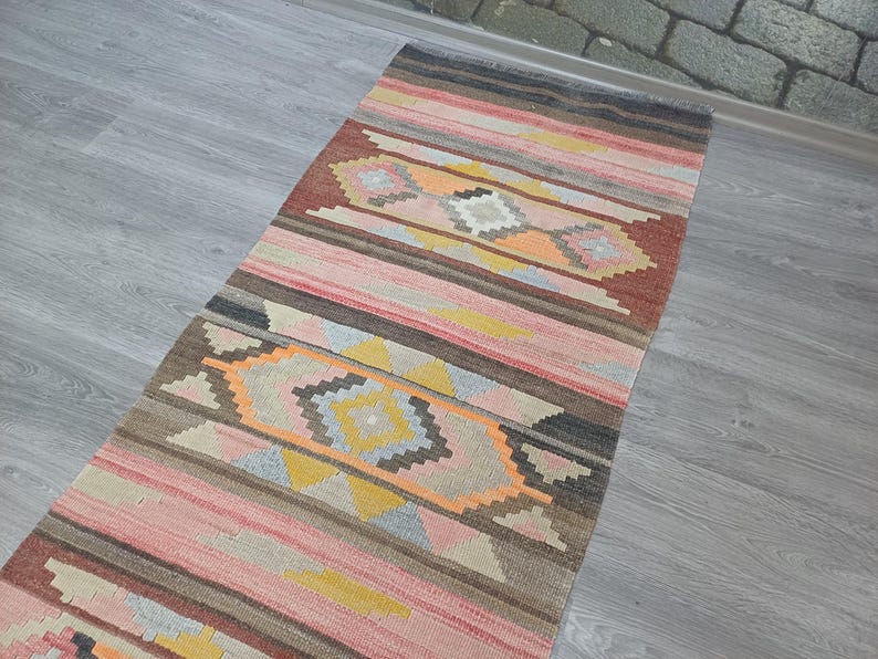 May include: A long, narrow rug with a colorful geometric pattern in shades of pink, orange, yellow, brown, and gray. The rug has a traditional, handwoven look.