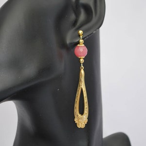 Puede incluir: Pendientes de aro dorados con una cuenta facetada rosa y un diseño de filigrana dorada.