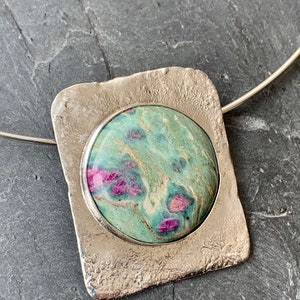May include: A silver pendant necklace featuring a green and pink gemstone set in a textured silver frame.