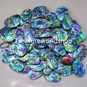 Op de afbeelding: Een grote groep iriserende abalone-schelpcabochons in verschillende tinten blauw, groen en paars. De cabochons zijn ovaalvormig en hebben een glad, gepolijst oppervlak.