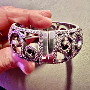 May include: A silver bangle bracelet featuring a delicate, intricate design. The bracelet is embellished with small, clear crystals that sparkle in the light.