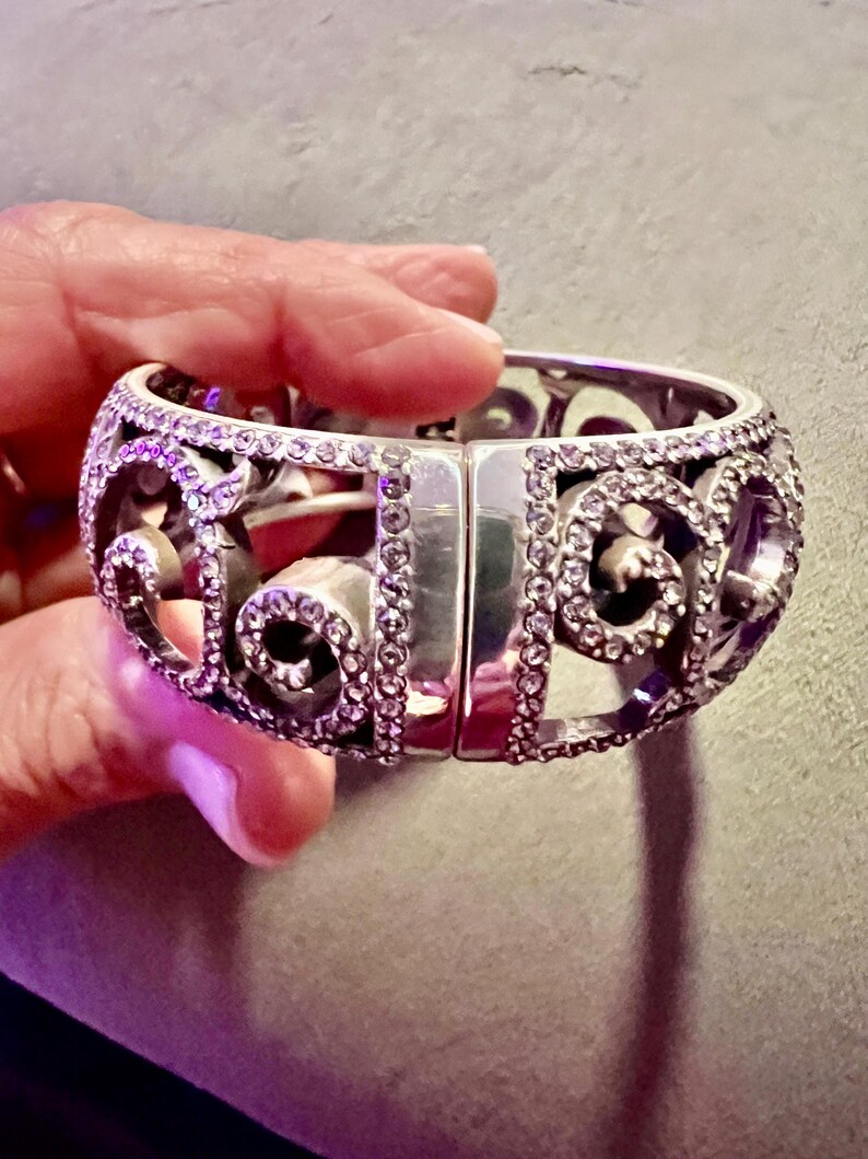 May include: A silver bangle bracelet featuring a delicate, intricate design. The bracelet is embellished with small, clear crystals that sparkle in the light.