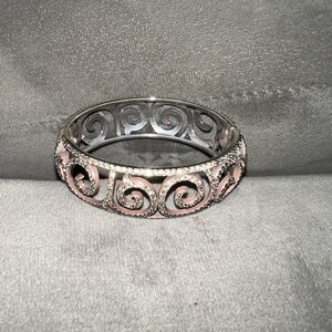 May include: A silver bangle bracelet with a pink enamel design featuring intricate swirls. The bracelet is made of metal and has a polished finish.