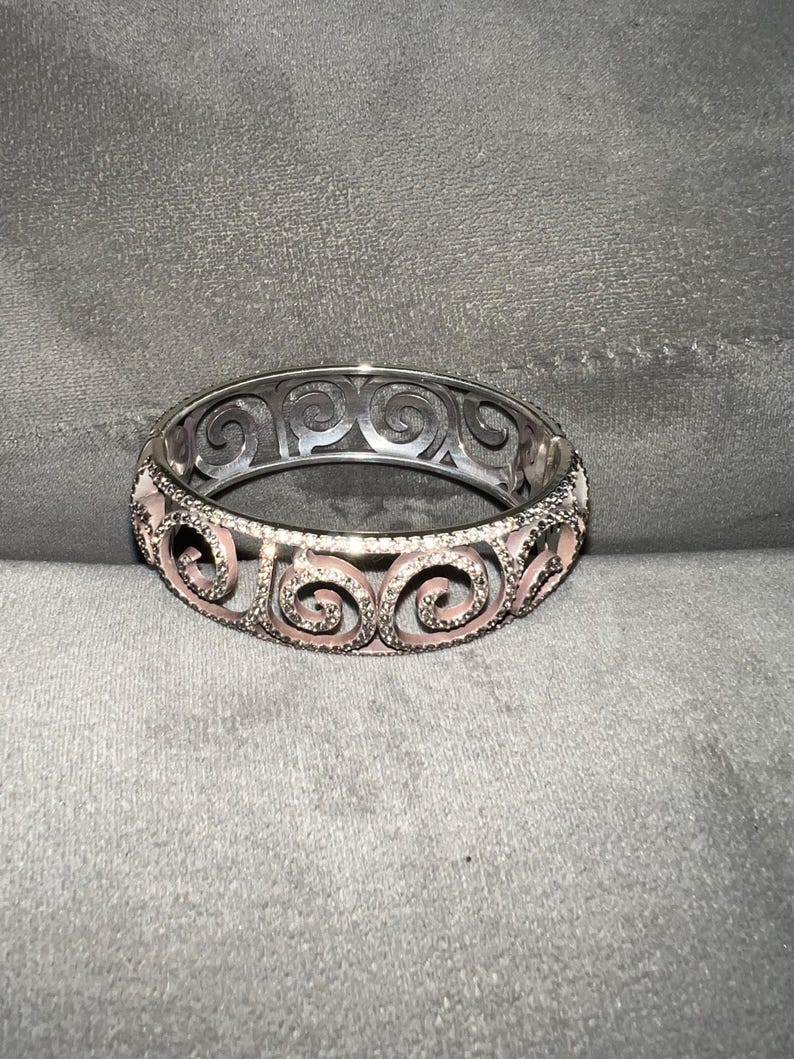 May include: A silver bangle bracelet with a pink enamel design featuring intricate swirls. The bracelet is made of metal and has a polished finish.