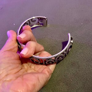 May include: A silver bangle bracelet featuring a delicate, intricate design. The bracelet is embellished with small, sparkling stones that create a shimmering effect.
