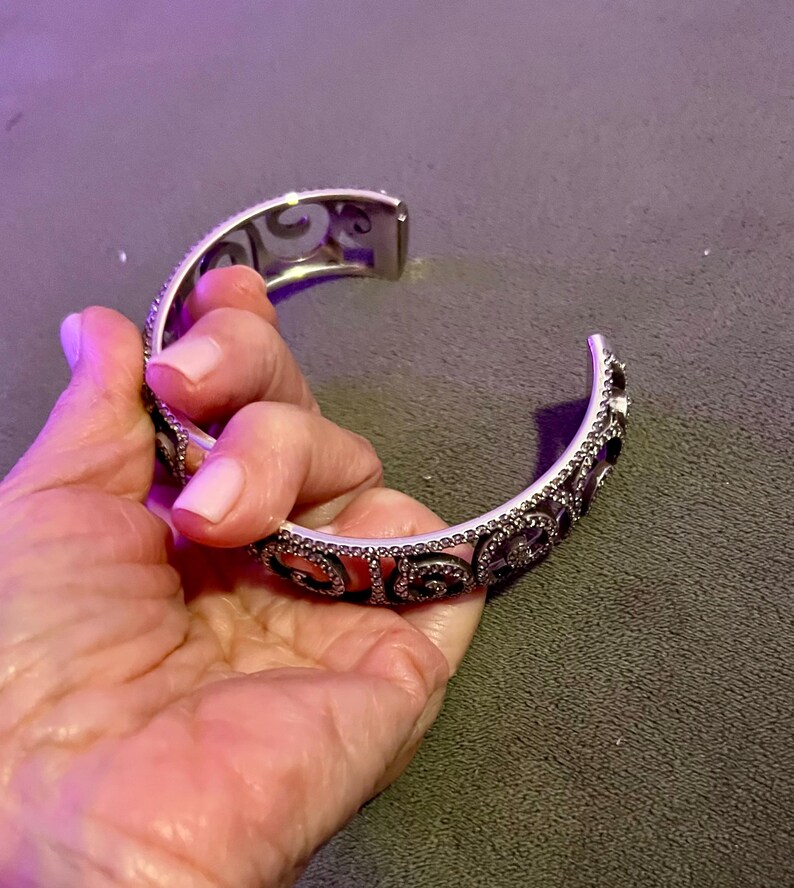 May include: A silver bangle bracelet featuring a delicate, intricate design. The bracelet is embellished with small, sparkling stones that create a shimmering effect.