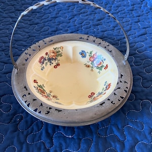May include: A vintage serving dish with a metal handle and rim. The dish has a cream-colored interior with floral designs in red, blue, and yellow. The metal rim features a decorative cut-out pattern.